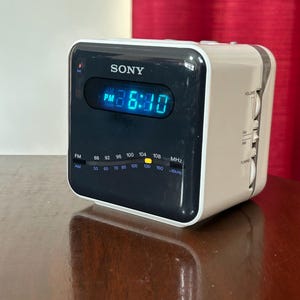 May include: A vintage Sony digital clock radio with a black face and a cream-colored body. The display shows the time as 6:40 PM in blue digits. It has FM and AM radio frequency indicators and volume and tuning knobs.
