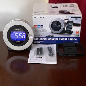 May include: A silver and black Sony Dream Machine CD clock radio with a circular digital display showing 5:56 in blue light. The set includes the original box, a manual, a small black remote control, and a black plastic docking adapter piece.