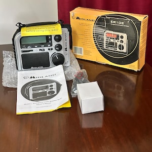 May include: A silver and black Midland ER102 emergency crank weather radio featuring a digital display, speaker, and control knobs. The set includes the original yellow and black product box, a white owner's manual, a small white box, and a plastic bag of parts.