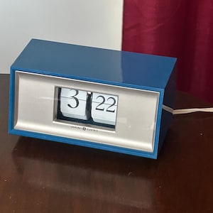 May include: A vintage blue General Electric flip clock displaying the time 3:22. The clock has a silver face and white numbers. The clock is rectangular and sits on a dark wooden surface.