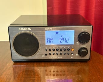 May include: A black Sangean WR-2 FM RDS/AM digital receiver with a brushed silver front panel. The device features a circular speaker grille, a blue backlit LCD screen displaying AM 1042, two large control knobs, and multiple small buttons.
