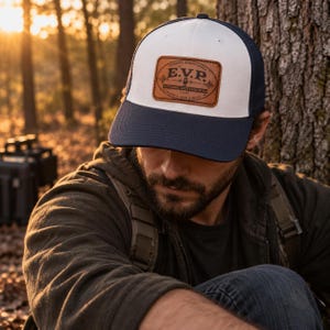 May include: A baseball cap with a white front panel, a dark blue brim, and a dark blue mesh back. A rectangular brown leather patch is centered on the front, featuring the text E.V.P. Electronic Voice Phenomena with a microphone icon.