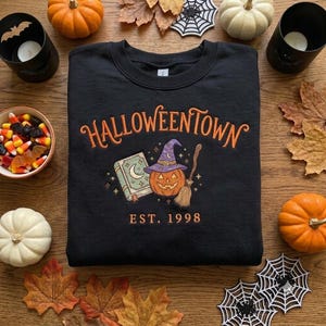 May include: A black crewneck sweatshirt featuring orange embroidered text that reads HALLOWEENTOWN EST. 1998. The design includes a light blue book with a crescent moon, an orange jack-o'-lantern wearing a purple witch hat, and a broomstick.