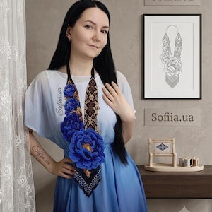 May include: A long beaded necklace featuring large, three-dimensional blue peony flowers with yellow centers, accented by intricate black and brown geometric beadwork patterns and a black fringe at the bottom, worn over a blue ombre dress.