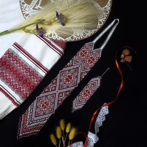Könnte beinhalten: Ein Set aus einer Krawatte und einem Armband aus Perlen mit einem geometrischen Muster in Rot, Schwarz und Weiß. Das Set wird zusammen mit einem weißen Stoff mit passender rot-schwarzer Stickkante, getrockneten Gräsern und weißer Spitze präsentiert.