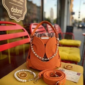Könnte beinhalten: Eine orangefarbene Beuteltasche aus gedrehtem Seil mit beigem Stoff-Kordelzug, einem goldfarbenen Kettenhenkel mit weißen Perlen und einem abnehmbaren, passenden orangefarbenen Seil-Schulterriemen. Ein weißes Perlenarmband liegt bei.