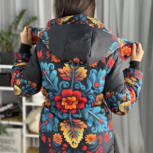 Peut inclure: Une veste à capuche noire ornée d'un motif floral multicolore vif dans des tons de rouge, orange, jaune et bleu. La veste comprend une taille à cordon de serrage, des manches longues avec des poignets noirs et une capuche assortie.