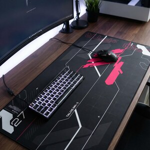 May include: A computer desk setup featuring a black and pink gaming mousepad with a white keyboard and a black mouse. A computer monitor, a black desk lamp, and a white computer case are also visible. The mousepad has the text "TALOS MK-1".