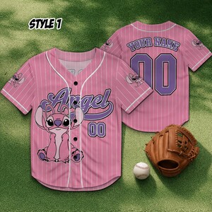 May include: Pink baseball jersey with white pinstripes, featuring the word "Angel" in purple script and a cartoon character. The jersey has the number "00" and a baseball glove and ball are next to it. The back of the jersey has the text "YOUR NAME" and the number "00".