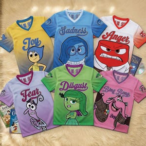 Könnte beinhalten: Sechs farbenfrohe Trikots zum Thema Alles steht Kopf. Jedes Trikot zeigt einen anderen Charakter und eine andere Emotion: Freude, Traurigkeit, Wut, Angst, Ekel und Bing Bong. Die Trikots haben einen V-Ausschnitt und werden auf einer flauschigen Oberfläche präsentiert.