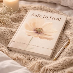 May include: A journal titled "Safe to Heal" with a floral design on the cover, alongside a pen with gold accents and a lit candle. The journal is open on a beige knitted blanket.
