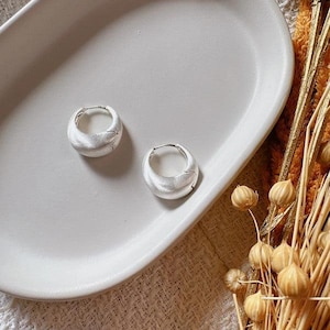 Peut inclure: Une paire de boucles d'oreilles créoles argentées avec une finition mate texturée. Les boucles d'oreilles sont présentées sur un plat ovale blanc, avec des accents floraux séchés en arrière-plan. Design arrondi et épais.