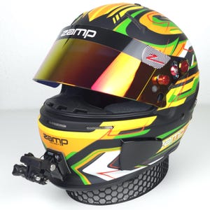 Puede incluir: Un casco de carreras Zamp con un diseño colorido con detalles en amarillo, verde y naranja sobre una base negra. El casco tiene una visera tintada y un soporte para cámara. El logotipo de Zamp es visible.