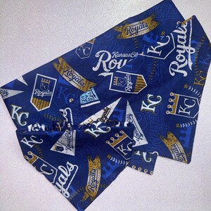 May include: Blue pet bandana with a repeating pattern of "Royals" text, "KC" logos, and baseball-themed graphics in white and gold. The bandana is folded and ready to wear.