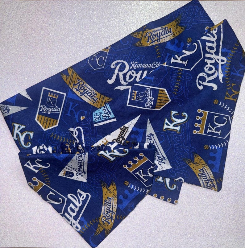 May include: Blue pet bandana with a repeating pattern of "Royals" text, "KC" logos, and baseball-themed graphics in white and gold. The bandana is folded and ready to wear.