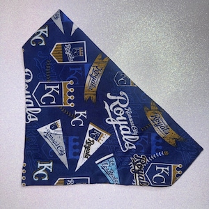 May include: A blue and white pet bandana featuring the Kansas City Royals baseball team logo and pennant designs. The bandana has the team's name and logo printed in white and gold.