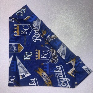 May include: A blue headband featuring a repeating pattern of the Kansas City Royals baseball team logo, including crowns, the letters "KC", and the word "Royals". The headband is made of fabric and has an elastic section.