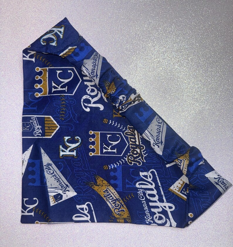 May include: A blue headband featuring a repeating pattern of the Kansas City Royals baseball team logo, including crowns, the letters "KC", and the word "Royals". The headband is made of fabric and has an elastic section.