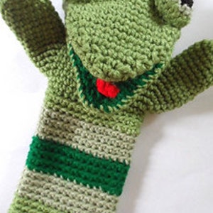 May include: Green crocheted frog puppet with a red tongue and black eyes. The puppet has a striped body with green, beige, and dark green stripes.
