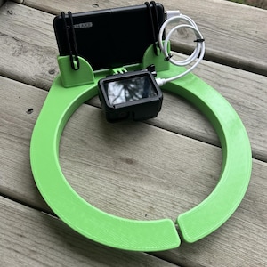 May include: A lime green, 3D-printed phone and camera holder. The holder has a circular design with a slot for a phone and a camera. A black phone and camera are secured in the holder with black elastic bands. A white charging cable is connected to the phone and camera.