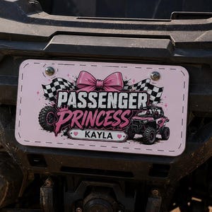 May include: A rectangular pink license plate attached to the rear of an off-road vehicle. The plate features a pink bow, checkered flags, and the text "PASSENGER PRINCESS" above the name "KAYLA" with small heart icons on each side of the name.