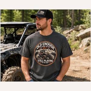 May include: A charcoal grey short-sleeve t-shirt featuring a circular graphic with a mountain landscape and an off-road vehicle. The text reads OLD ENOUGH TO KNOW BETTER BUT STILL PLAYING IN THE DIRT. A black baseball cap with a small rectangular patch.