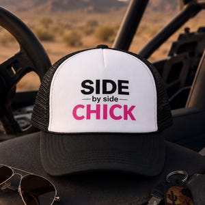 Könnte beinhalten: Eine schwarz-weiße Trucker-Cap mit weißer Vorderseite, auf der der schwarze Schriftzug SIDE, der kleinere schwarze Text -by side- und der große pinke Schriftzug CHICK gedruckt sind. Die Cap hat ein schwarzes Netzteil und einen schwarzen Schirm.