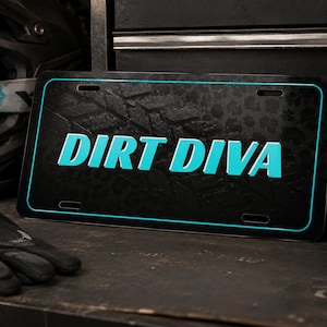 May include: A rectangular black license plate featuring the text DIRT DIVA in bold cyan lettering. The plate has a thin cyan border and a subtle dark tire tread pattern across the background. It includes four mounting slots at the corners.