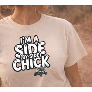 Könnte beinhalten: Cremefarbenes T-Shirt mit dem Text "I'M A SIDE (BY SIDE) CHICK" in fetter, umrandeter Schrift. Unter dem Text befindet sich eine kleine Grafik eines Side-by-Side-Fahrzeugs. Das T-Shirt ist ein Freizeitartikel.