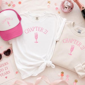 May include: A selection of birthday-themed items, including a white t-shirt, a beige sweatshirt, and a pink trucker hat, each featuring the text "CHAPTER 21" and a champagne glass design. Also, heart-shaped sunglasses and a card that says "CHEERS TO 21 YEARS".