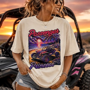 Könnte beinhalten: Ein cremefarbenes Kurzarm-T-Shirt mit einem Grafikdruck eines Dünenbuggys an einem Strand bei Sonnenuntergang. Der Text "Passenger Princess" ist in einer stilisierten Schriftart über und unter der Illustration gedruckt, die Palmen und Wolken zeigt.