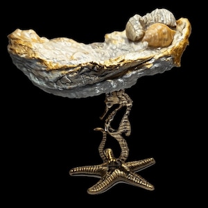 May include: A decorative oyster shell-shaped dish with gold trim, holding small seashells. The dish is supported by a seahorse and starfish base, all in a metallic finish. The design is set against a black background.