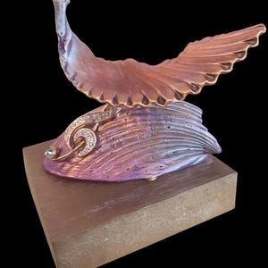 Op de afbeelding: Een bronzen sculptuur van een vogel met uitgespreide vleugels, geplaatst op een paars en gouden voet. De voet staat op een rechthoekig, bruin blok. De sculptuur heeft een decoratief, met juwelen bezet accent.