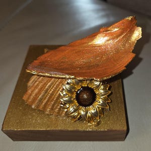 Pode incluir: Uma peça decorativa artesanal com um girassol dourado, uma concha e um elemento em forma de folha. A folha e a concha são pintadas em tons de laranja e dourado, montadas numa base de madeira dourada.