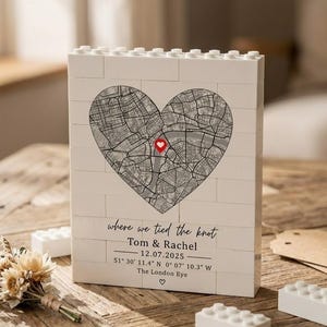 Puede incluir: Un bloque blanco estilo ladrillo con un mapa en forma de corazón y un marcador rojo. El texto incluye "where we tied the knot", nombres, una fecha y coordenadas. Un pequeño ramo y bloques blancos están sobre una superficie de madera.