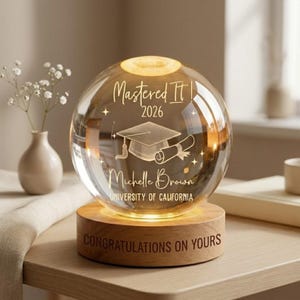 May include: A clear glass globe with a graduation cap and scroll design, engraved with "Mastered It 2026" and a name. The globe sits on a wooden base with the words "Congratulations on Yours". A decorative graduation gift.