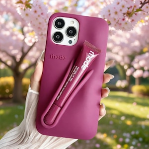 May include: A magenta phone case with the brand name "rhode" printed on it. A matching magenta lip tint tube is attached to the case. The lip tint tube has the word "mode" printed on it.