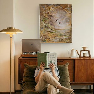 May include: A rectangular framed wall art piece featuring an abstract landscape painting with two hummingbirds in flight. The artwork displays textured brushstrokes in shades of brown, gold, green, and white, enclosed within a thin black frame.