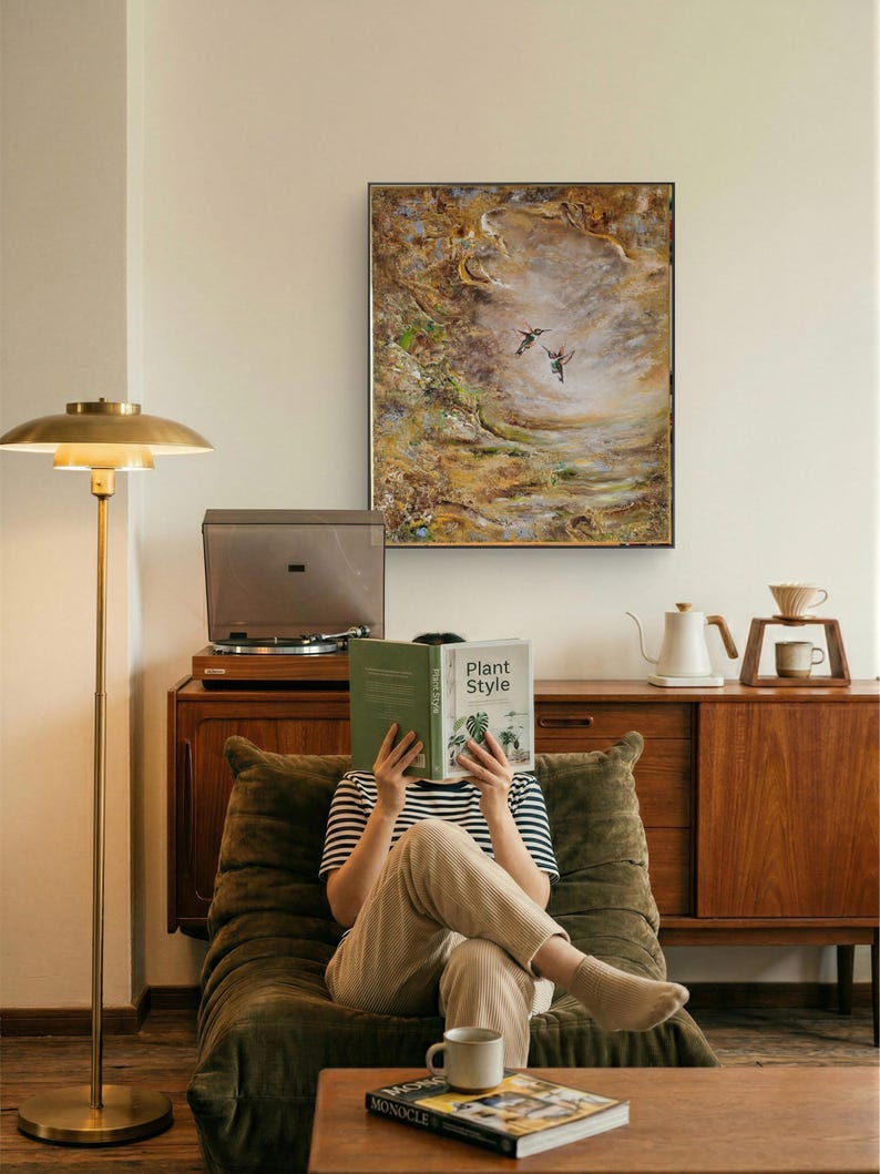 May include: A rectangular framed wall art piece featuring an abstract landscape painting with two hummingbirds in flight. The artwork displays textured brushstrokes in shades of brown, gold, green, and white, enclosed within a thin black frame.