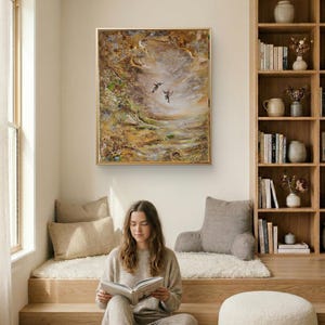 May include: A rectangular framed painting featuring an abstract landscape in shades of gold, brown, and cream with two small birds flying in the center. The artwork is displayed on a plain wall above a seating area with cushions and a bookshelf.