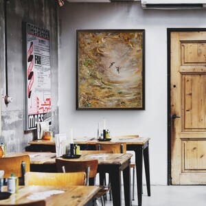 May include: A rectangular framed painting featuring two small birds in flight against a textured, abstract background of earthy brown, gold, and muted green tones, displayed on a white wall above wooden tables in a dining space.