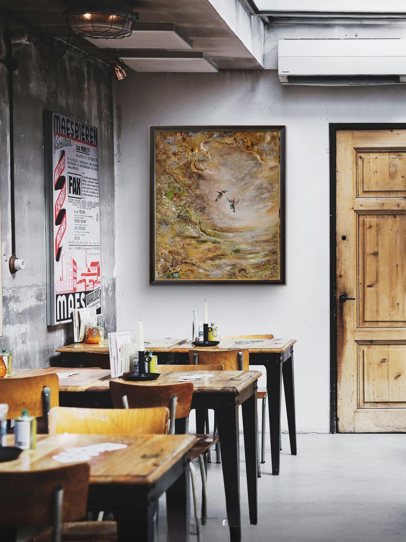 May include: A rectangular framed painting featuring two small birds in flight against a textured, abstract background of earthy brown, gold, and muted green tones, displayed on a white wall above wooden tables in a dining space.