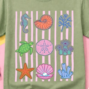 Puede incluir: Camiseta verde salvia con un gráfico colorido de vida marina. El diseño incluye un caballito de mar, una concha de nautilus, coral, una tortuga, un dólar de arena, una estrella de mar, una almeja con una perla y otros elementos marinos. La camiseta tiene rayas verticales rosas.