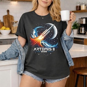 Puede incluir: Camiseta gris oscuro con un gráfico colorido de un cohete lanzándose hacia una luna, con el texto "ARTEMIS II 2026". El diseño incluye elementos azules, naranjas y blancos, creando una ilustración de temática espacial.