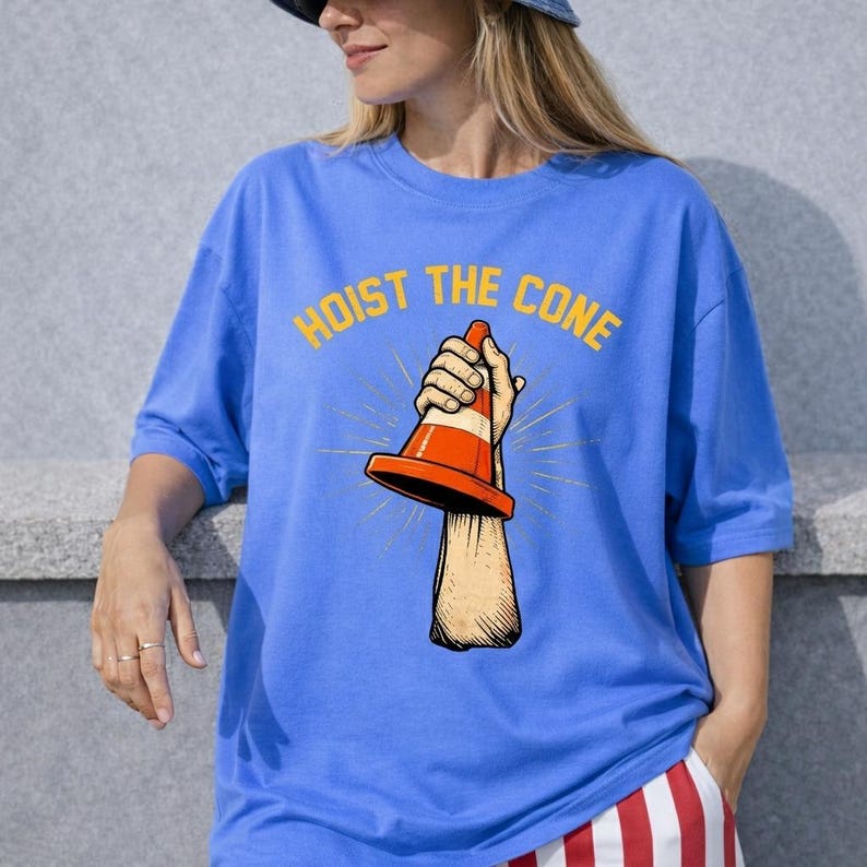 May include: A blue t-shirt featuring the text "HOIST THE CONE" in yellow. The graphic depicts a hand holding an orange and white traffic cone. The shirt is a solid blue colour.