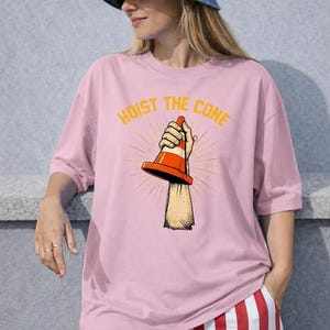 May include: Pale pink t-shirt with the text "HOIST THE CONE" in yellow. The graphic shows a hand holding an orange and white traffic cone. The shirt is a plain pink colour.