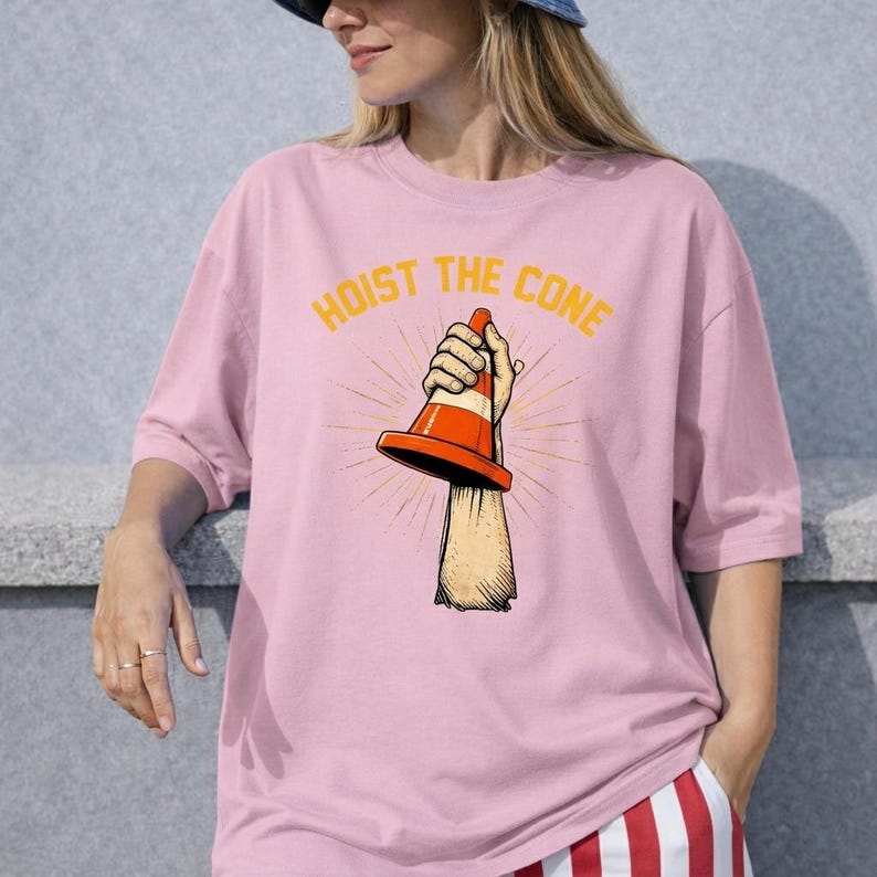 May include: Pale pink t-shirt with the text "HOIST THE CONE" in yellow. The graphic shows a hand holding an orange and white traffic cone. The shirt is a plain pink colour.