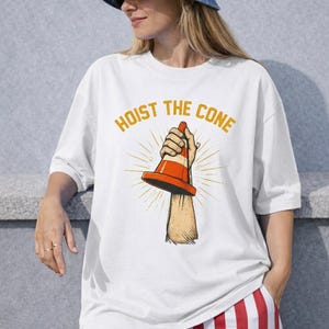 May include: White t-shirt with the text "HOIST THE CONE" in gold lettering. The graphic features a hand holding an orange and white traffic cone, with sunburst rays emanating from behind the cone.