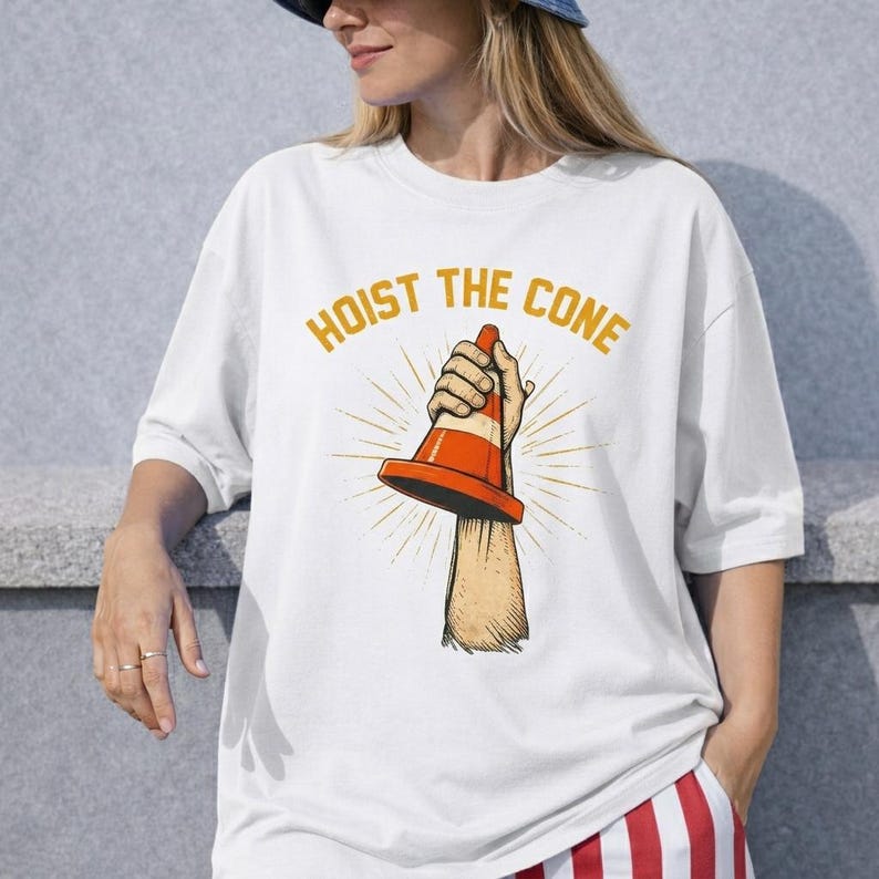 May include: White t-shirt with the text "HOIST THE CONE" in gold lettering. The graphic features a hand holding an orange and white traffic cone, with sunburst rays emanating from behind the cone.