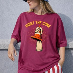 May include: A maroon t-shirt with the text "HOIST THE CONE" in yellow. The graphic shows a hand holding an orange and white traffic cone. The shirt is worn by a person with a denim hat and striped trousers.
